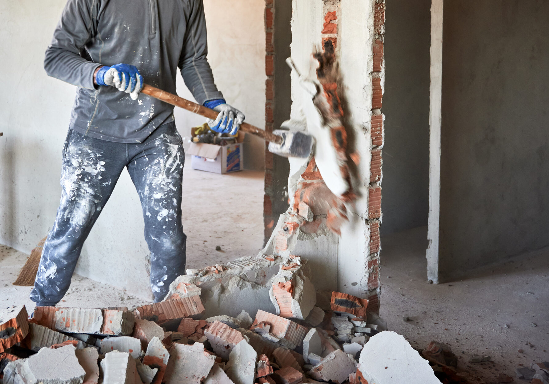 Close up of sledgehammer blow on brick, plaster. Workman striking devastating blow at remnants of interoom wall against backdrop of table and other plastered walls.